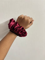 Hand wearing a large burgundy satin scrunchie on a plain background