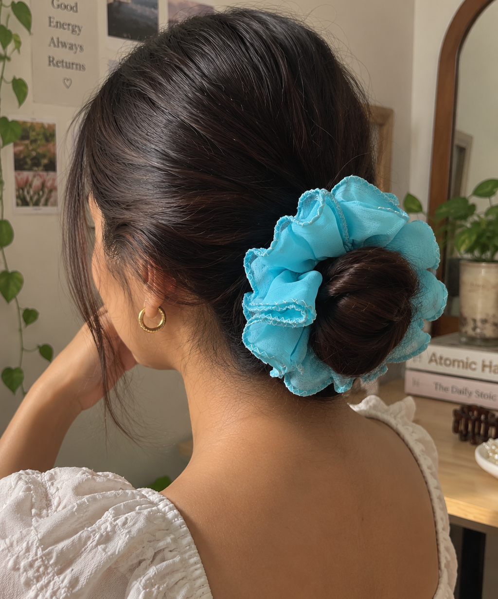 A woman shown from the back wearing a light blue ruffled scrunchie hair tie in her dark hair, set against an indoor background with a mirror, plants, and wall art.