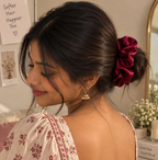 A woman with dark hair styled in a low bun wearing a deep burgundy satin scrunchie, shown from the back and side. She is wearing a cream-colored floral top with red patterns and gold earrings in a warm indoor setting.
