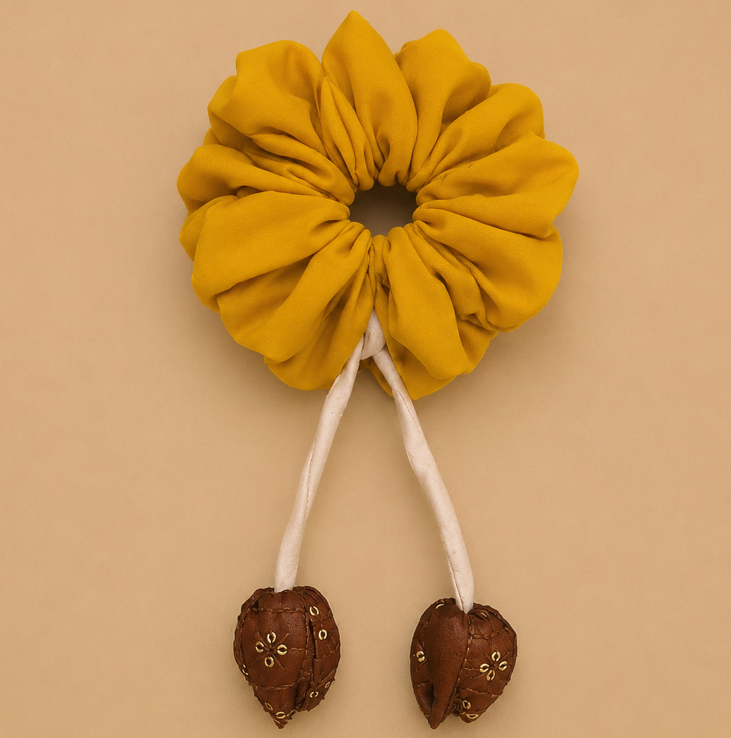 A mustard yellow fabric scrunchie with two long white hanging cords ending in brown heart-shaped beads, displayed on a solid beige background.